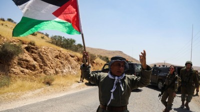 مواطن فلسطيني يحمل علم فلسطين وخلفه جنود إسرائيليين (رويترز)